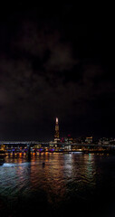 Fototapeta premium The River Thames illuminated by the reflections of the colored lights of the buildings and skyscrapers at night in London with the cloudy night sky with bridges crossing and illuminating the river.