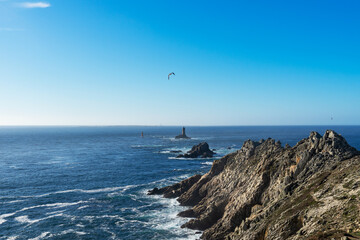 La Pointe du Raz, avec le Phare de la Vieille sur son îlot rocheux, tandis que l'île de Sein se profile à l'horizon, un paysage emblématique de la côte bretonne. - obrazy, fototapety, plakaty