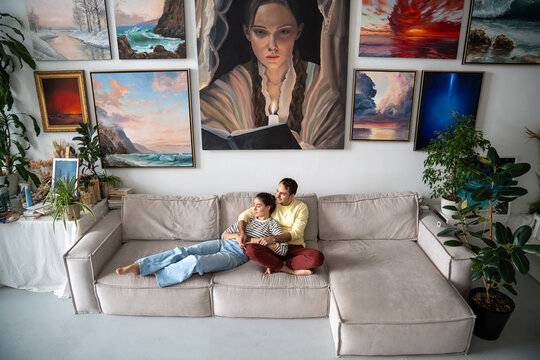 Happy couple hugging, dreaming, resting on sofa after a working day in spacious bright loft room of gallery. In interior of art studio composition of many paintings on wall. Slow life concept.  - Powered by Adobe
