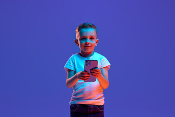 Child, schoolboy in casual attire using smartphone in mixed neon light against blue background. Internet safety for children. Concept of fashion and style, beauty, back to school, e-learning. Ad