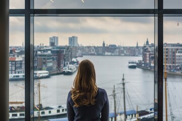 Fototapeta na wymiar A woman looks outside a large window of an office building overlooking the harbor