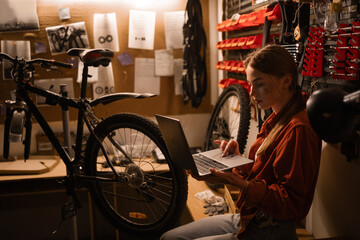Female repairman doing bike servicing in workshop or repair shop, looking on laptop maintenance list.