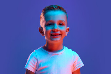 Portrait of charming child, boy dressed white T-shirt looking at camera widely smiling in mixed neon light against blue background. Concept of human emotions, fashion and style, beauty, back to school