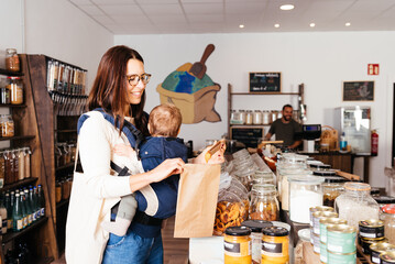 Woman Holding Baby in Carrier Shopping in Bulk Grocery Store