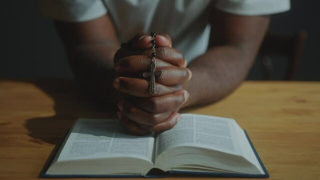 Hands of unrecognizable African American man holding rosary beads with crucifix and leaning on wooden table with opened Bible during prayer. Close-up view