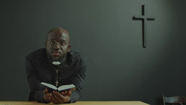 Black Catholic priest in cassock leaning on altar in modern church with cross on the wall, holding Bible, looking upwards with hope and saying prayer to God