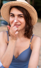 Young woman in straw hat and blue swimsuit applying sun screen cream on her face on the beach.