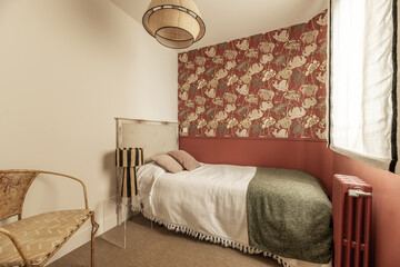 Bedroom with decorative wallpaper on the walls, single bed with wooden headboard upholstered in fabric, window with blinds, metal armchair with upholstery, red cast iron radiator