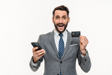Happy Caucasian young businessman showing credit card and using cellphone isolated over white...
