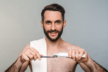 Hygienic procedures concept. Sexy young shirtless man brushing teeth isolated over grey background. Handsome Caucasian male model holding tooth brush and paste looking at the camera
