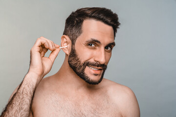 Funny young man using cotton stick swab to clean ears isolated over grey background. Male beauty concept, cleaning, morning routine, healthcare, hygienic everyday procedures