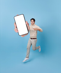 Full length of cheerfull Asian man jumping and smiling in air with showing mobile phone blank...