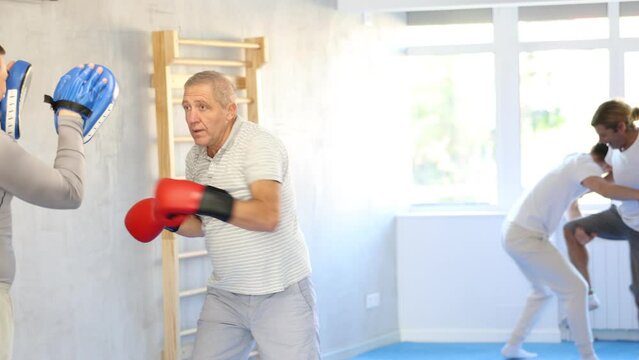Coach and mature man pupil are engaged during training, boxing classes. Male trainer holds makiwara and helps student to work out force of blow, to box quickly and effectively. High quality 4k footage