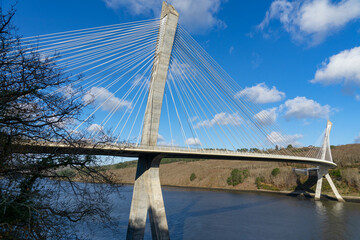 Sous un ciel bleu éclatant, le pont de Térénez, pont à haubans courbé au-dessus de l'Aulne, offre une vue emblématique à l'entrée de la presqu'île de Crozon, dans le Finistère, en Bretagne. - obrazy, fototapety, plakaty