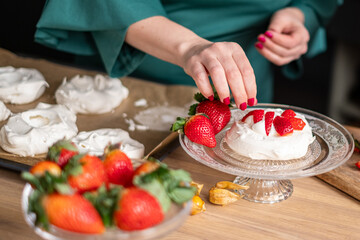 A chef in a green apron assembles strawberry pavlovas in a bright kitchen, with more desserts in...