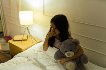 Young latin girl with her teddy bear, feeling sick and coughing while trying to sleep in her bedroom at home