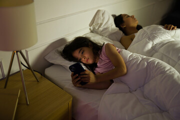 Little girl stays awake at night while using a smartphone to play some games and check on social media in their softly lit bedroom