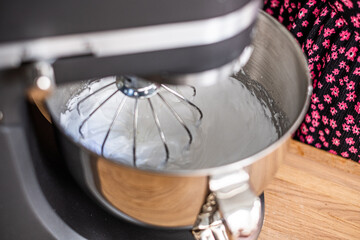 Close up of a stand mixer whipping meringue, with a glimpse of a baker in a floral shirt in the...