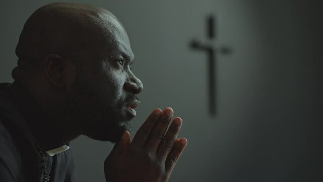 African American priest wearing cassock and clerical collar holding palms together and praying to God in church. Close-up shot, side view
