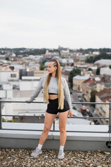 A young woman stands on a rooftop dressed in stylish athletic wear, overlooking a panoramic cityscape. Her relaxed posture and thoughtful expression reflect a moment of peace amidst urban life.