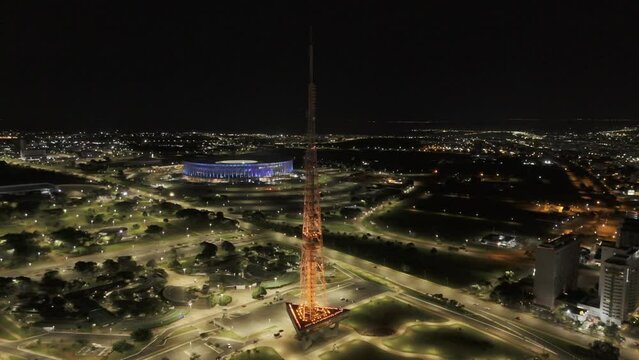 Drone orbits around orange TV Tower as it changes to red, with blue Arena BRB Mane Garrincha in background and lit up Ferris Wheel in Castelinho do Parque da Cidade in Brasilia, Brazil