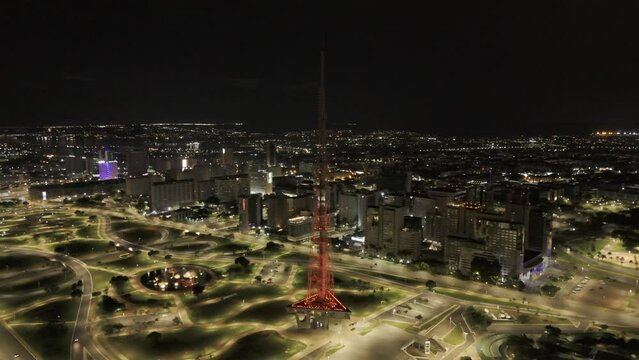 Drone orbits around red TV Tower as it changes to green while overlooking Jardim Burle Marx at night in Brasilia, Brazil