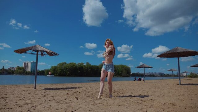 Woman dancing bachata, salsa and kizomba on the beach against the background of the blue sky. Smiling Woman barefoot poses and dances seductively on hot sand. Beach scene with girl enjoying the moment
