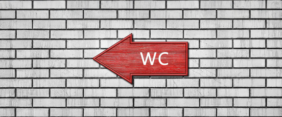 Red wooden arrow sign with the inscription WC hanging on a white brick wall. Left arrow pointer