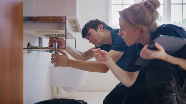 Female plumber holding digital tablet with male trainee fixing leaking pipe in bathroom sink - shot in slow motion