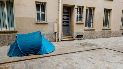 Blue camping tent pitched on a cobblestone urban street, symbolizing homelessness and poverty, with...