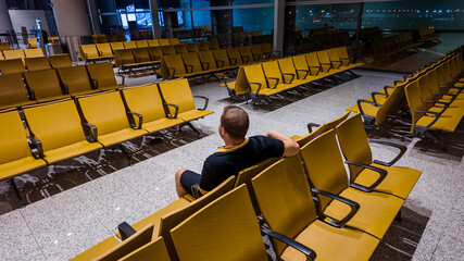 Adult man sitting alone amidst rows of empty chairs in a well-lit airport waiting area, depicting...