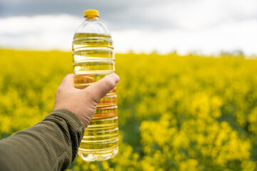 A bottle of rapeseed oil in a hand against the background of a yellow blooming rapeseed field. A...