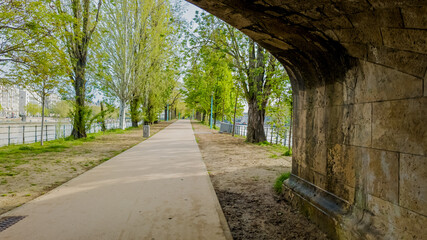 Tranquil riverside promenade with tall green trees in spring, under an old stone bridge arch, ideal for concepts related to leisure and urban nature