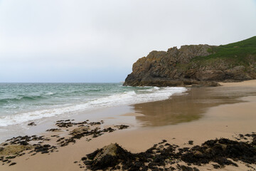 Falaise abrupte, plage déserte, sable mouillé : la Bretagne se dévoile sous un ciel nuageux, mêlant mystère et beauté brute.