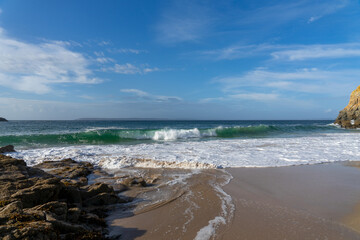 Belle vague vert émeraude et écume blanche dans la mer d'Iroise, bordée par une plage de sable et des rochers, illustrant la diversité côtière de la Bretagne.