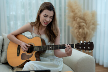 A woman enjoying a relaxed moment, playing guitar while sitting on a couch at home, surrounded by a...