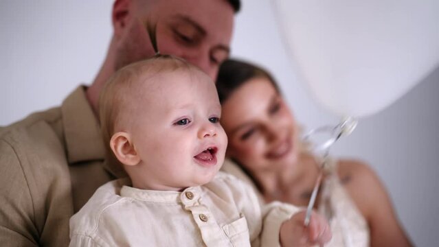 Happy little baby holding a balloon. Loving parents at backdrop in blur. Family portrait in studio.