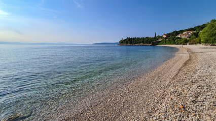 A stony beach along the shore of Medveja in Croatia. The Mediterranean Sea is calm and clear. There is a lush forest with a small town at the shore. Clear, blue sky. Summer remedy. Holidays vibes.