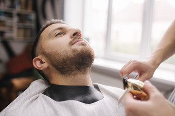 Professional hairdresser uses a hair clipper for fringing beard for a handsome man in barbershop....