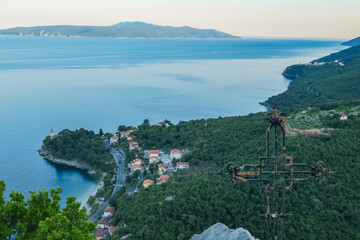 A small metal cross on a rock with a panoramic view of the shore along Medveja, Croatia seen from...