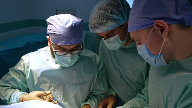 Doctor holds his hand up waiting for the instrument. Assisting staff gives the tool to a surgeon and trauma surgery goes on. Three doctors lean to the operated area.