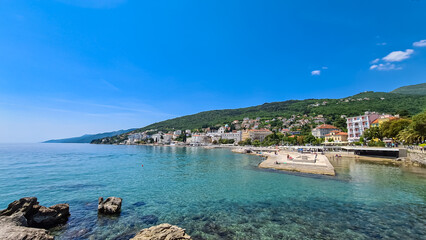 A panoramic view of the shore along Opatija, Croatia. There is a town located on the shore of the Mediterranean Sea. Calm surface of the sea. Stony beach. Green hills in the back. Sunny day. Cloudless