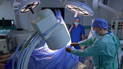 C-Arm x-ray machine is set over the patient covered with blue sheet at operation. Medic arrange the equipment in surgical room. - Powered by Adobe