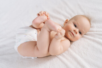 Portrait of sweet beautiful newborn baby in diapers lying on bedsheets, plays touches toes smiling looking at camera. Carefree healthy babyhood, healthcare and pediatrics care concept.