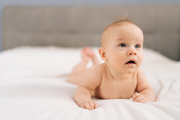 Closeup of cute little baby girl lying on stomach on bed, learning to hold head. Portrait of nice newborn naked baby looking interestingly away with open mouth. Carefree healthy babyhood concept.