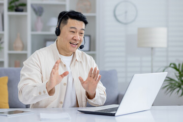 A joyful older Asian man wearing a headset and interacting cheerfully with someone on his laptop...