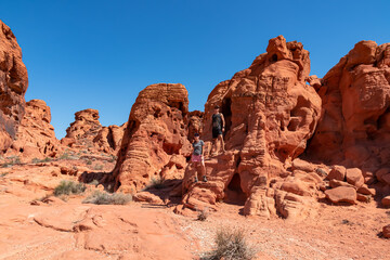 Fototapeta premium Couple at the entrance of windstone arch (fire cave) in Valley of Fire State Park, Mojave desert, Nevada, USA. Scenic view of beehive shaped red sandstone rock formations. Barren deserted landscape