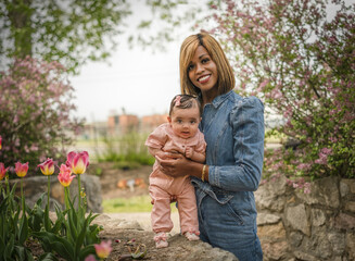 Portrait of beautiful African American woman with her adorable baby girl in park with blooming flowers in spring