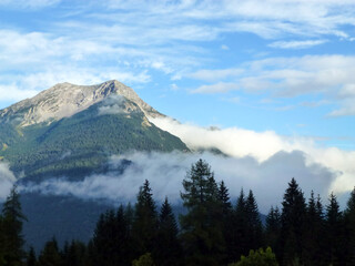 Obraz premium A picturesque view of a mountain rock overgrown with a green forest. Above them is a blue sky and white clouds