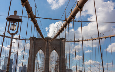 The gate of Brooklyn Bridge with a waving American flag on top of it contrasted with a blue sky with puffy, white clouds. Suspension bridge in New York City. Numerous skyscrapers in the back.